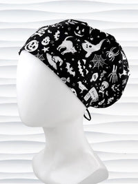 A black fabric Euro Pixie style surgical scrub cap by JensCaps with a white Halloween-themed print of ghosts, bats, spiders, and jack-o'-lanterns, displayed on a white mannequin head 