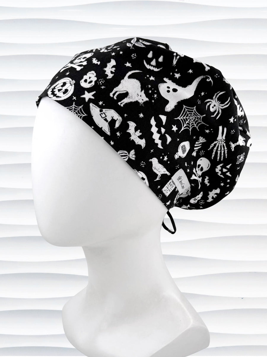 A black fabric Euro Pixie style surgical scrub cap by JensCaps with a white Halloween-themed print of ghosts, bats, spiders, and jack-o'-lanterns, displayed on a white mannequin head 