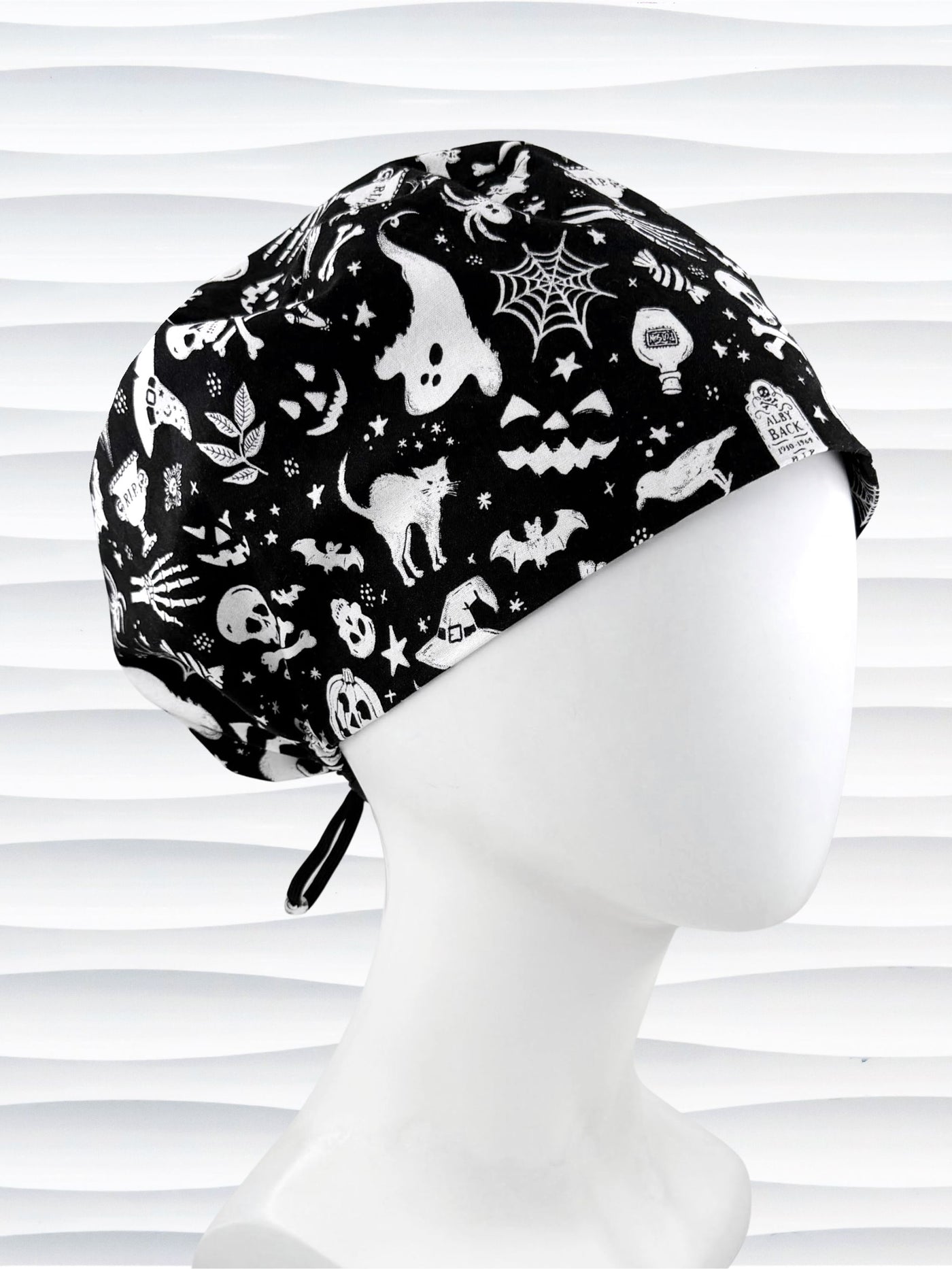 A black and white Halloween-themed scrub cap with a pattern of ghosts, bats, spiders, and pumpkins, displayed on a white mannequin head against a white wavy background.