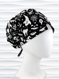 A black and white Halloween-themed scrub cap with a pattern of ghosts, bats, spiders, and pumpkins, displayed on a white mannequin head against a white wavy background.