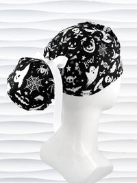 Black and white Halloween-themed scrub cap by JensCaps displayed on a white mannequin head. The fabric features a spooky print of bats, ghosts, skulls, and pumpkins, accented with a white ribbon bow.