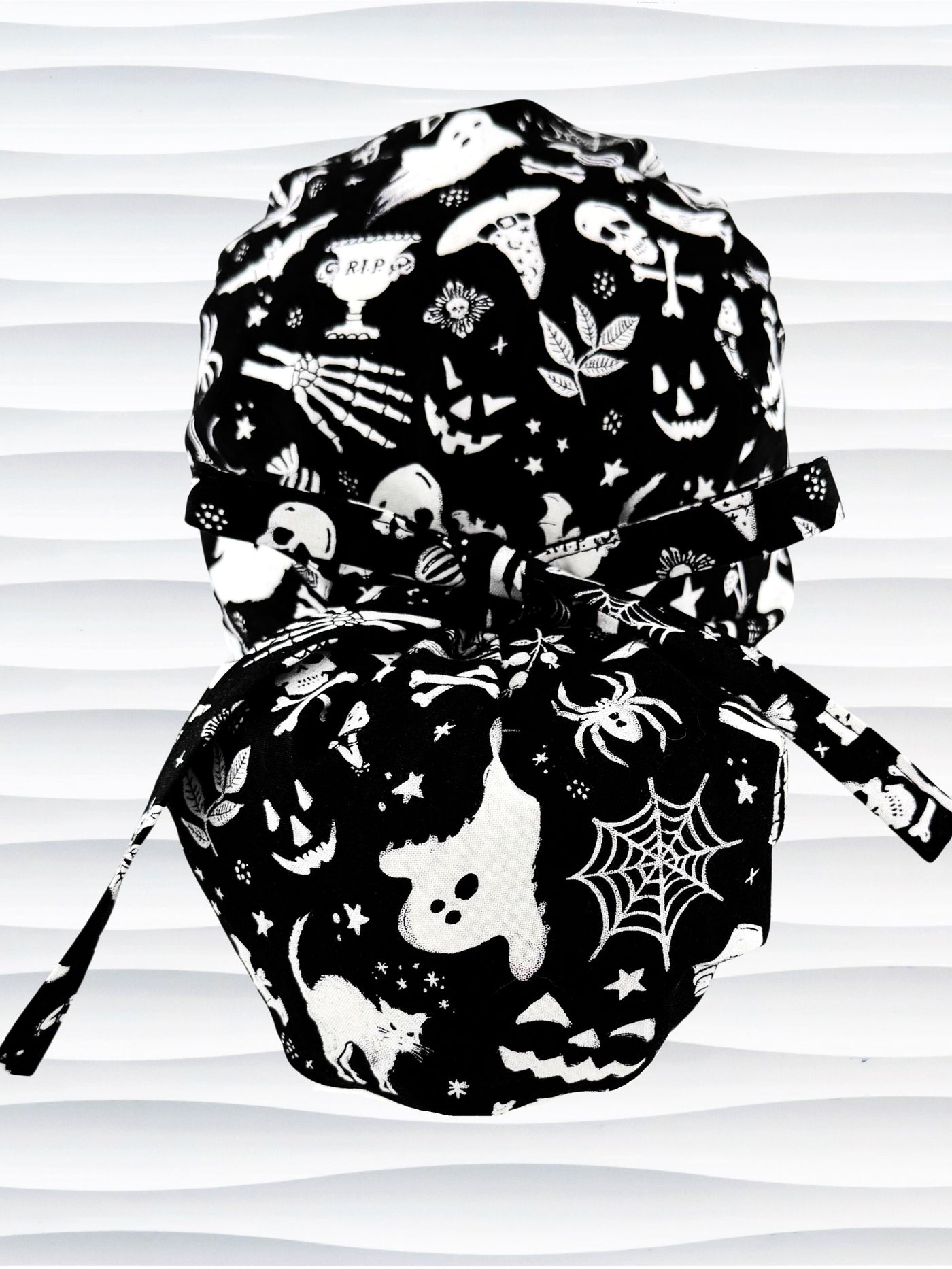 Ponytail style Black and white Halloween-themed scrub cap by JensCaps featuring spooky motifs like ghosts, spiders, and jack-o'-lanterns, displayed on a white mannequin head and shown against a wavy white background.