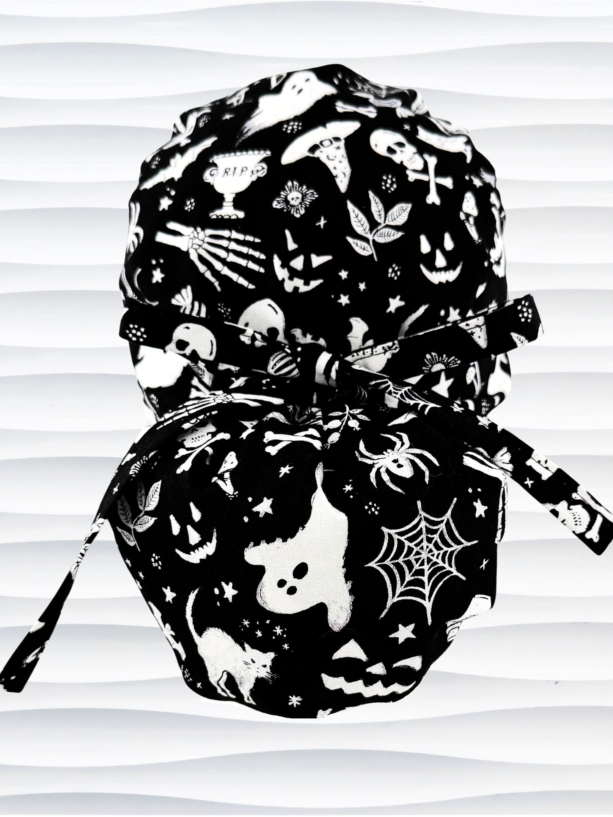 Black and white Halloween-themed Euro Pony style scrub cap by JensCaps with spiderwebs, bats, skulls, and ghost patterns, displayed on a white mannequin head with a fabric bow.