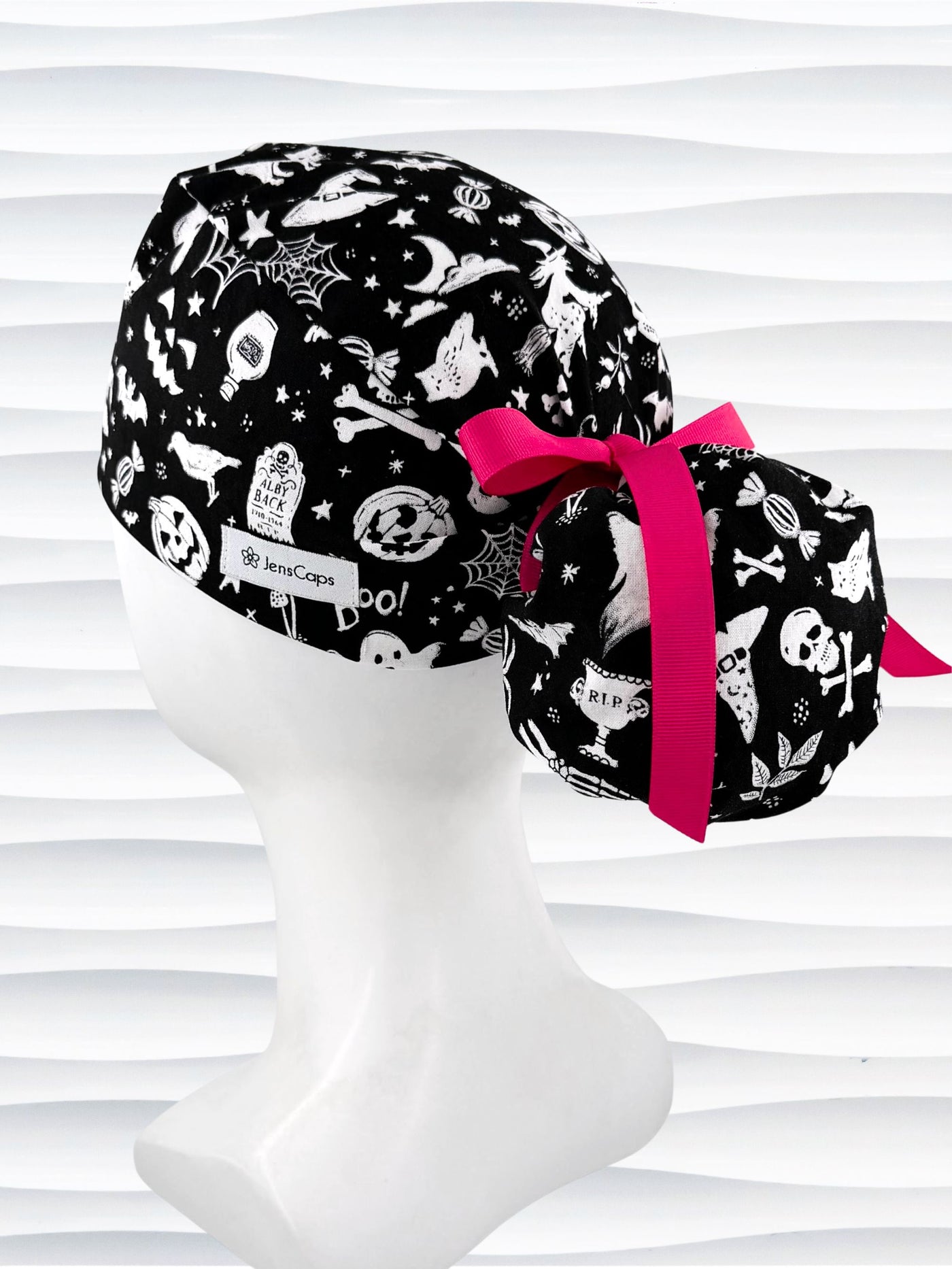 Black and white Halloween-themed scrub cap by JensCaps displayed on a white mannequin head. The fabric features a spooky print of bats, ghosts, skulls, and pumpkins, accented with a bright pink ribbon bow.