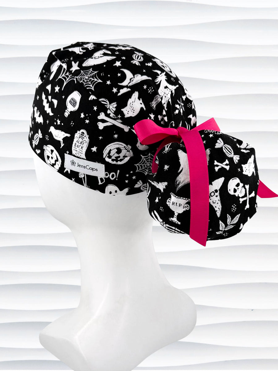 Black and white Halloween-themed scrub cap by JensCaps displayed on a white mannequin head. The fabric features a spooky print of bats, ghosts, skulls, and pumpkins, accented with a bright pink ribbon bow.
