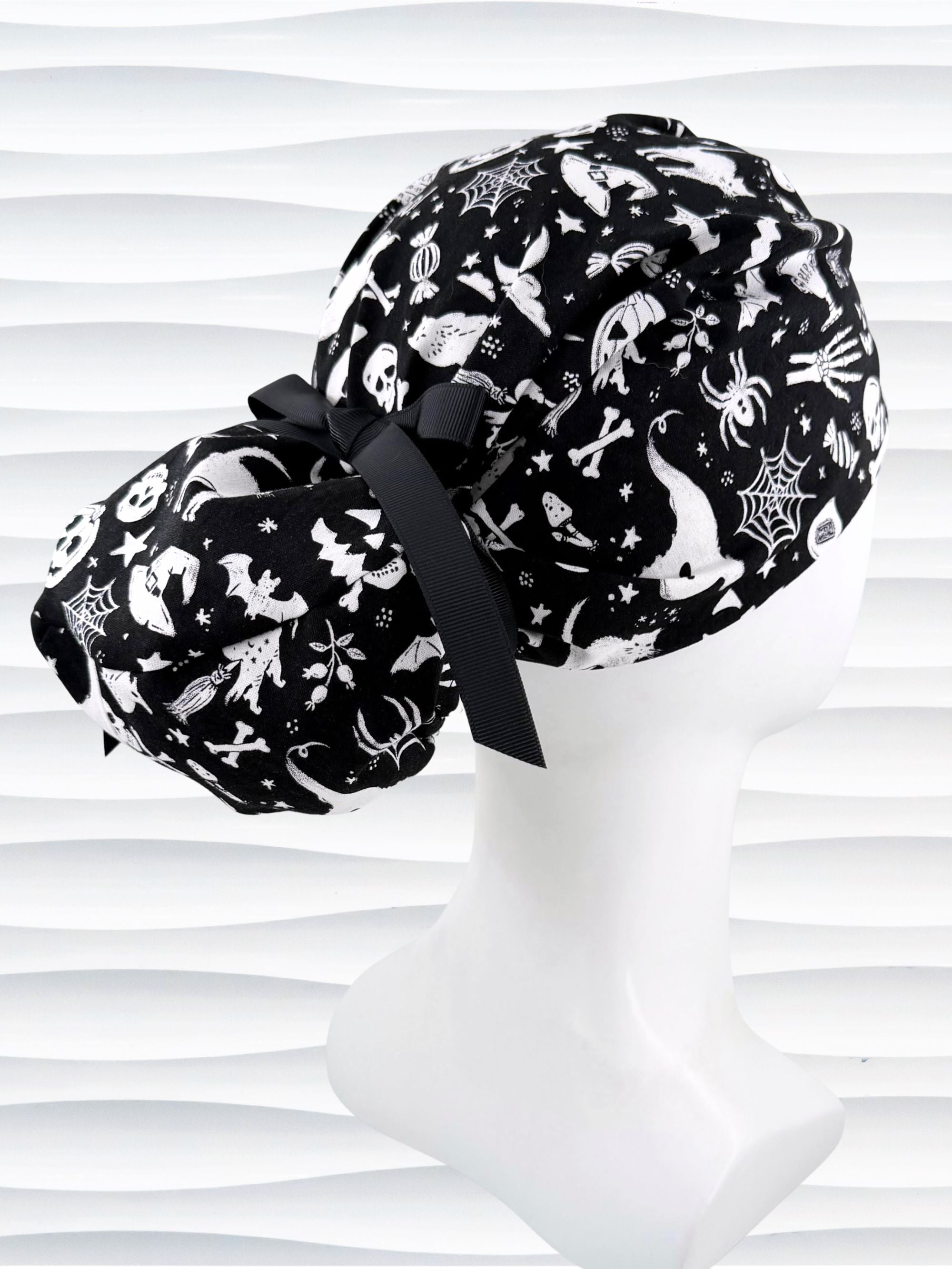 Black and white Halloween-themed Euro Pony style scrub cap by JensCaps with spiderwebs, bats, skulls, and ghost patterns, displayed on a white mannequin head with a black ribbon bow.