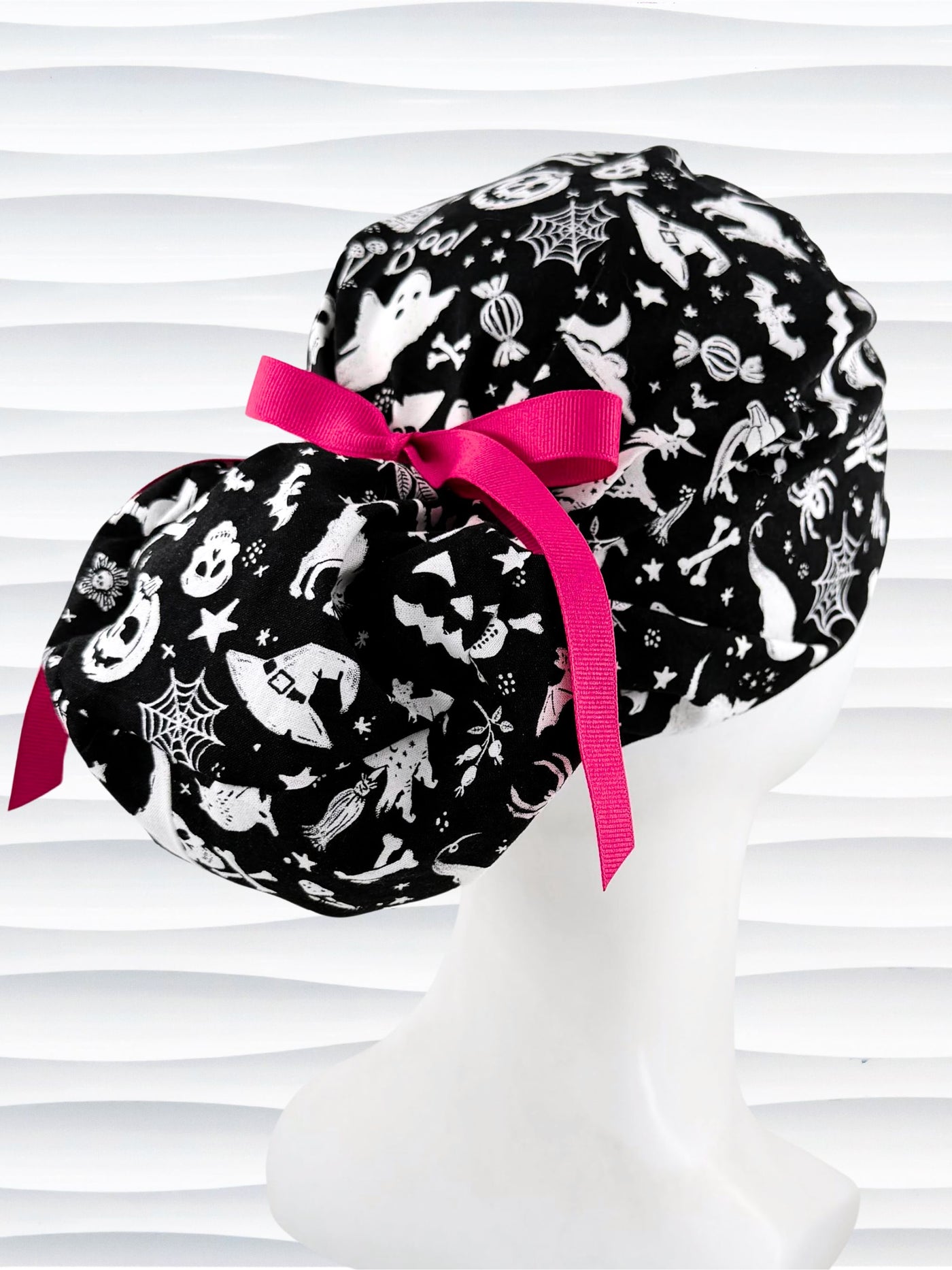 Black and white Halloween-themed Euro Pony style scrub cap by JensCaps with spiderwebs, bats, skulls, and ghost patterns, displayed on a white mannequin head with a pink ribbon bow.