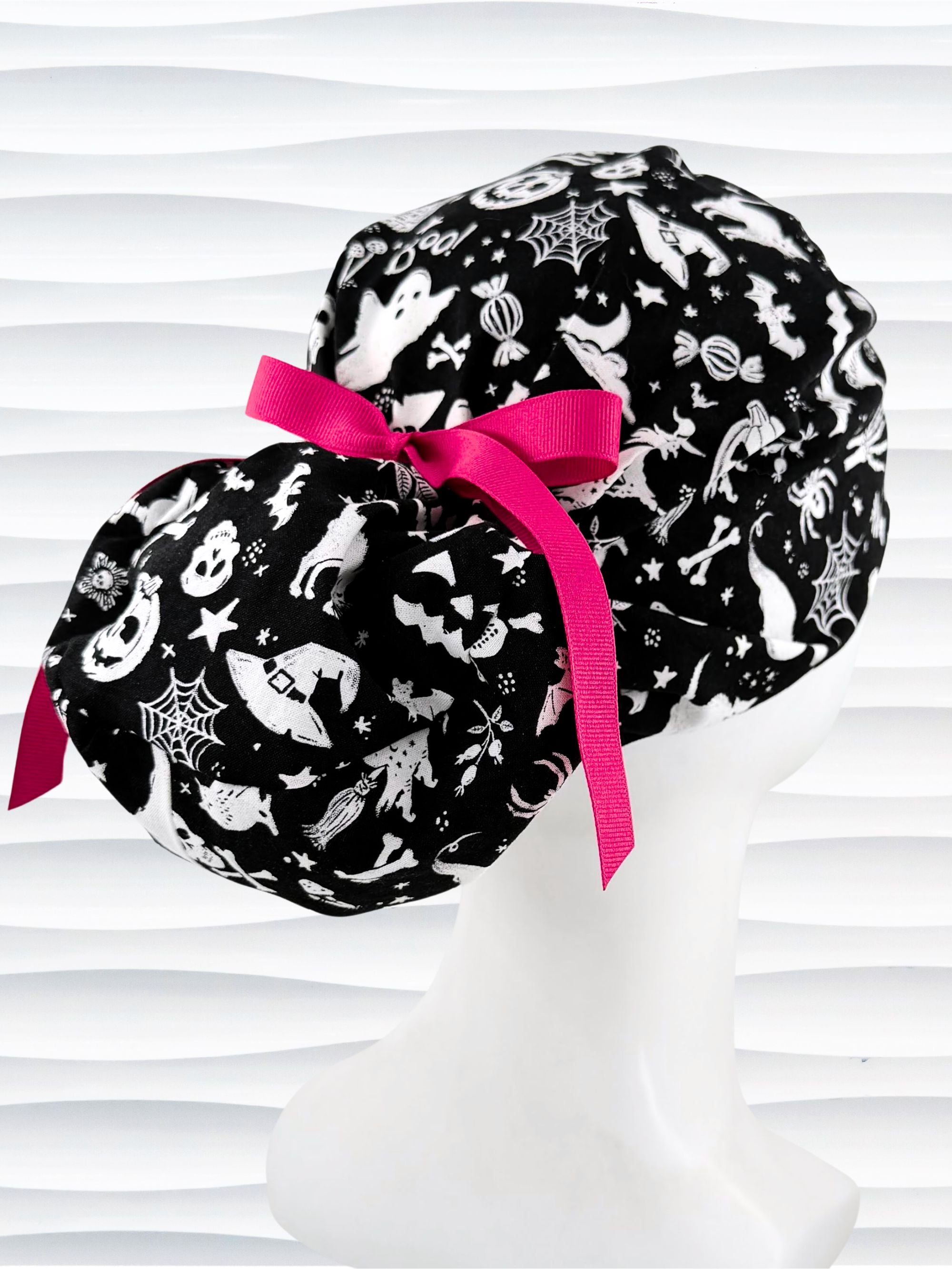 Black and white Halloween-themed Euro Pony style scrub cap by JensCaps with spiderwebs, bats, skulls, and ghost patterns, displayed on a white mannequin head with a pink ribbon bow.
