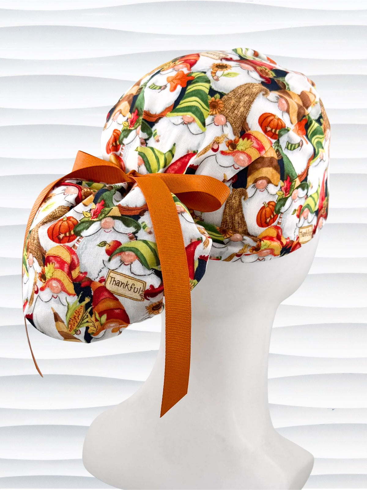 Ponytail style surgical scrub cap with gnomes in fall colors holding sunflowers and signs that say thankful, tied with a ribbon, and displayed on a mannequin head.