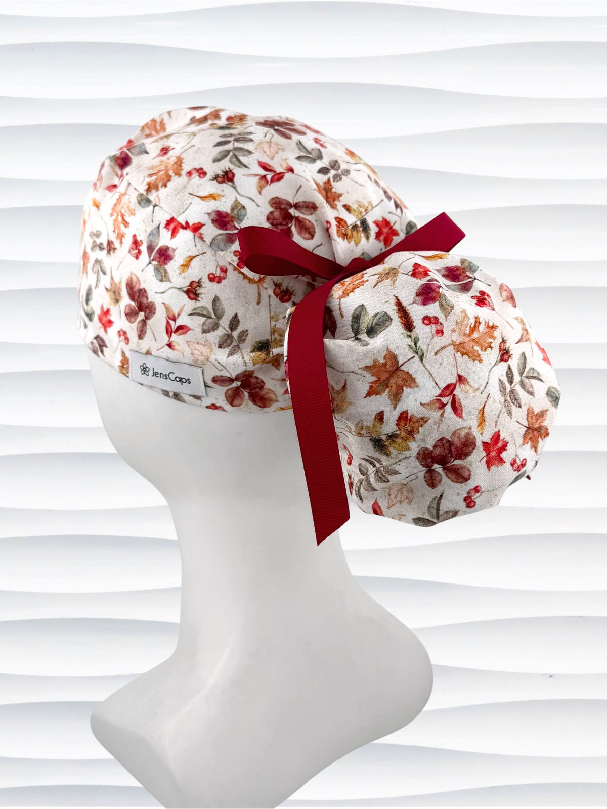 Ponytail style surgical scrub cap with an assortment of autumn eaves in fall colors all over a light background, finished with a ribbon tie, and displayed on a white mannequin head.