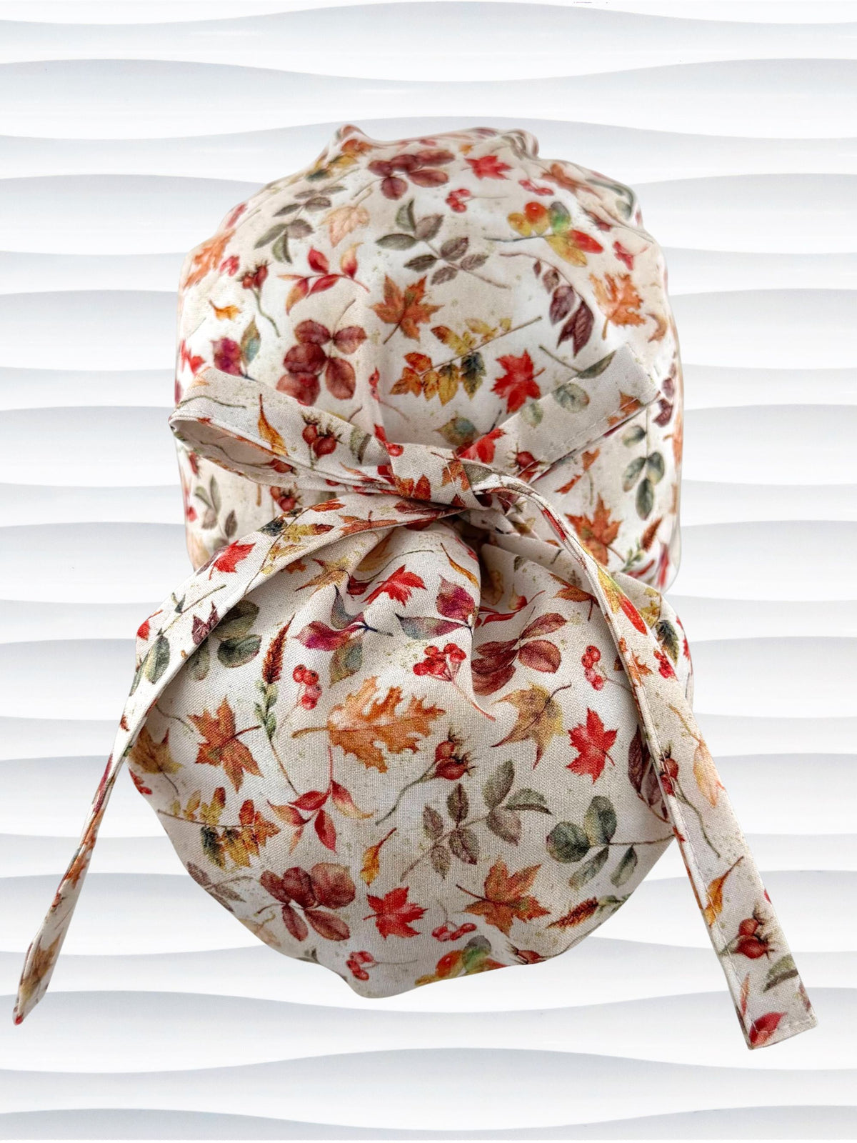 Euro Pony style surgical scrub cap with an assortment of autumn eaves in fall colors all over a light background, finished with a ribbon tie, and displayed on a white mannequin head.