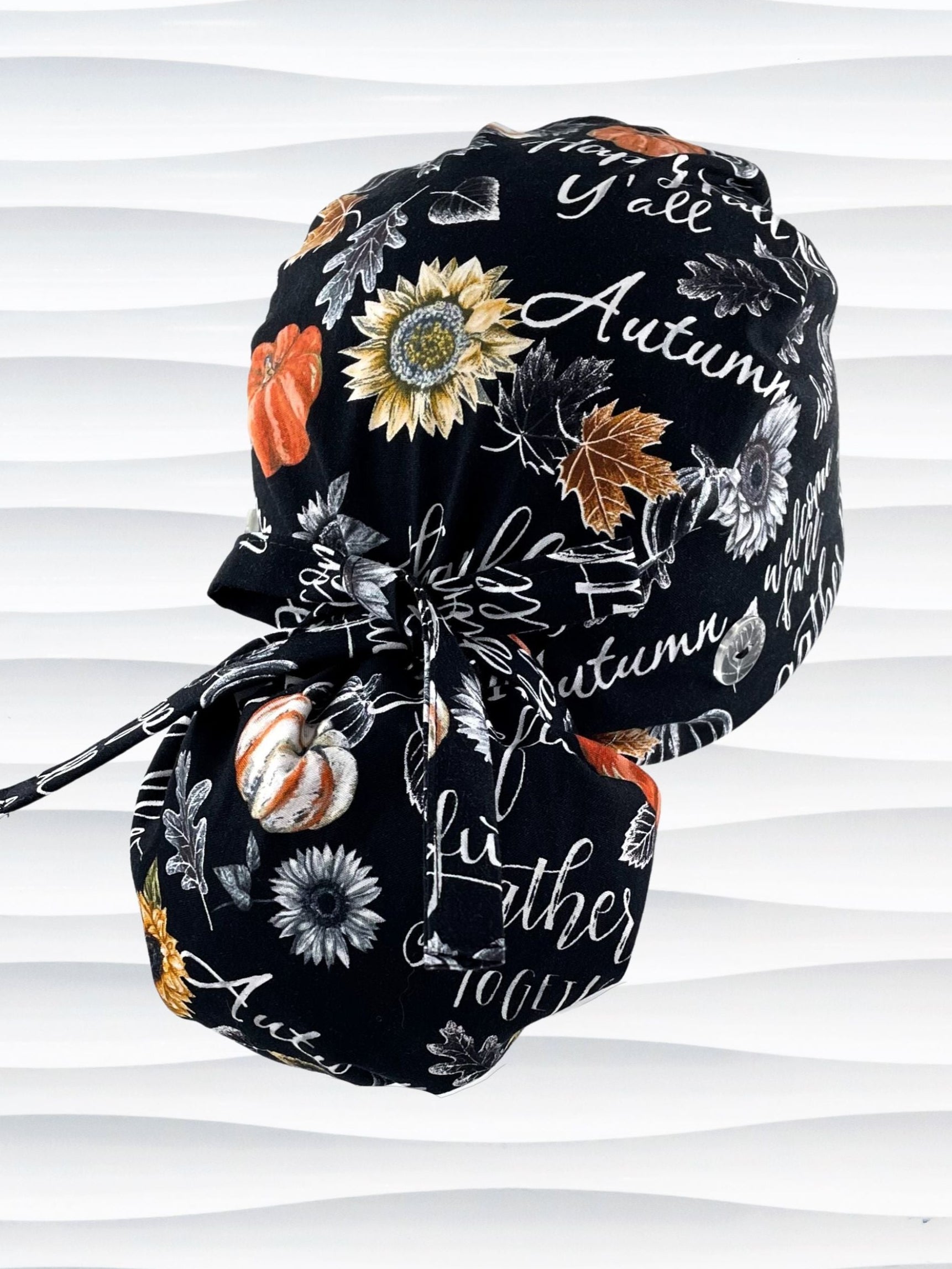Ponytail style surgical scrub cap hat for Thanksgiving and Autumn with "Happy Fall Y'all " and other holiday script and sunflowers on black cotton fabric.