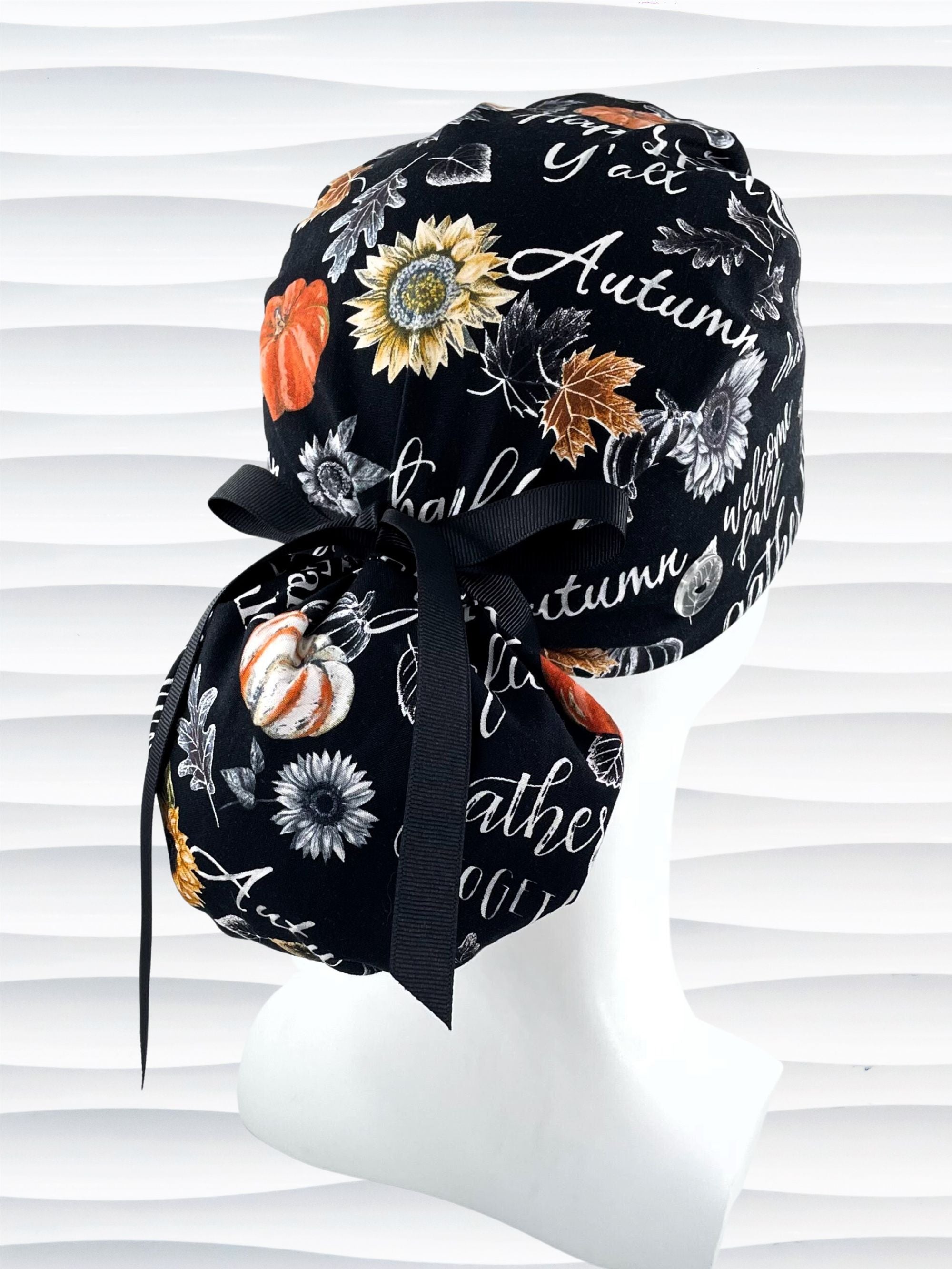 Ponytail style surgical scrub cap hat with pumpkins, sunflowers, leaves, and autumn themed words all over black cotton fabric.
