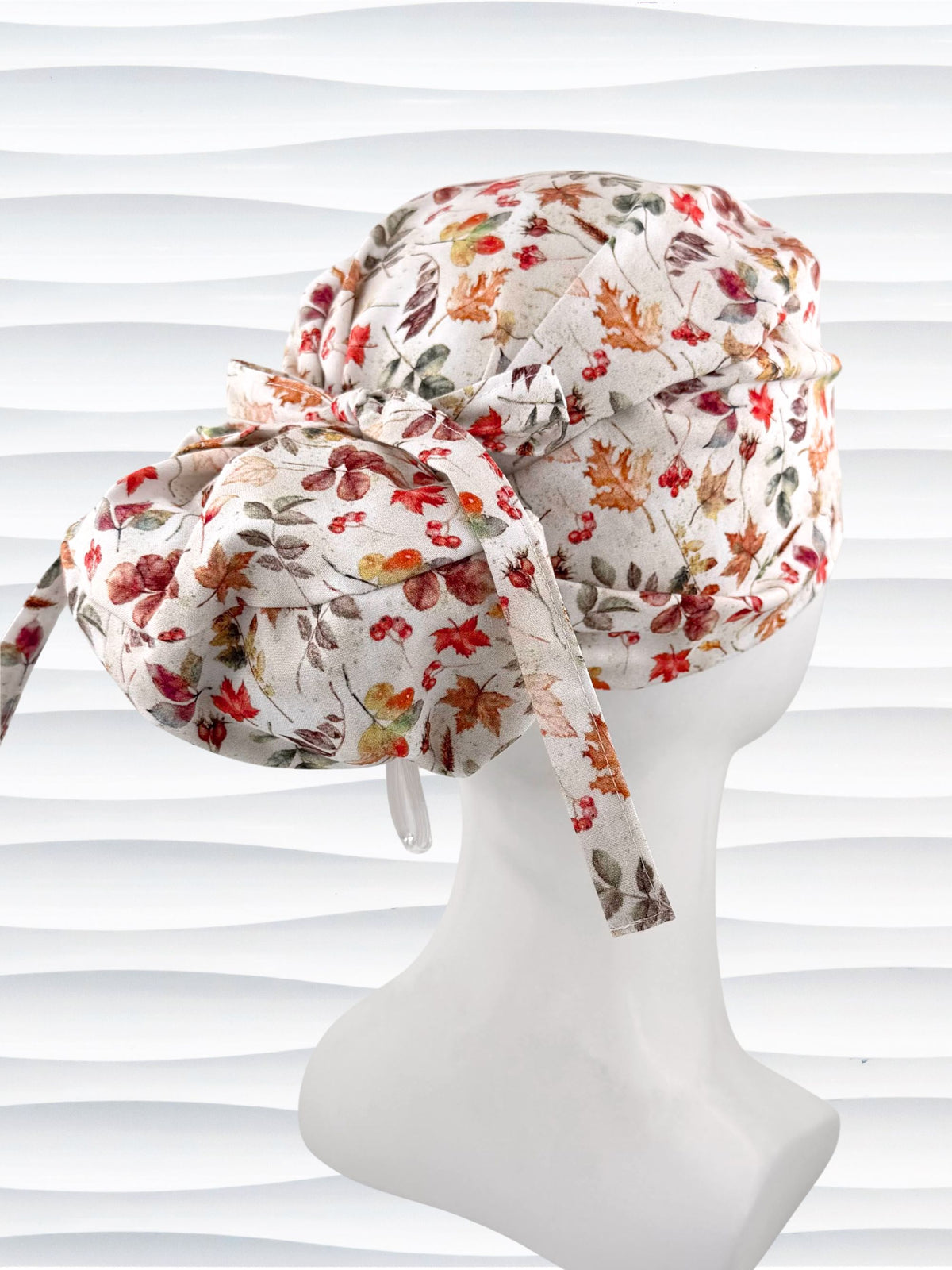 Euro Pony style surgical scrub cap with an assortment of autumn eaves in fall colors all over a light background, finished with a ribbon tie, and displayed on a white mannequin head.