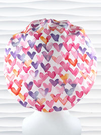 Bouffant style surgical scrub cap by JensCaps with watercolor hearts in pastel colors all over white cotton fabric and dispoayed on a white mannequin head.