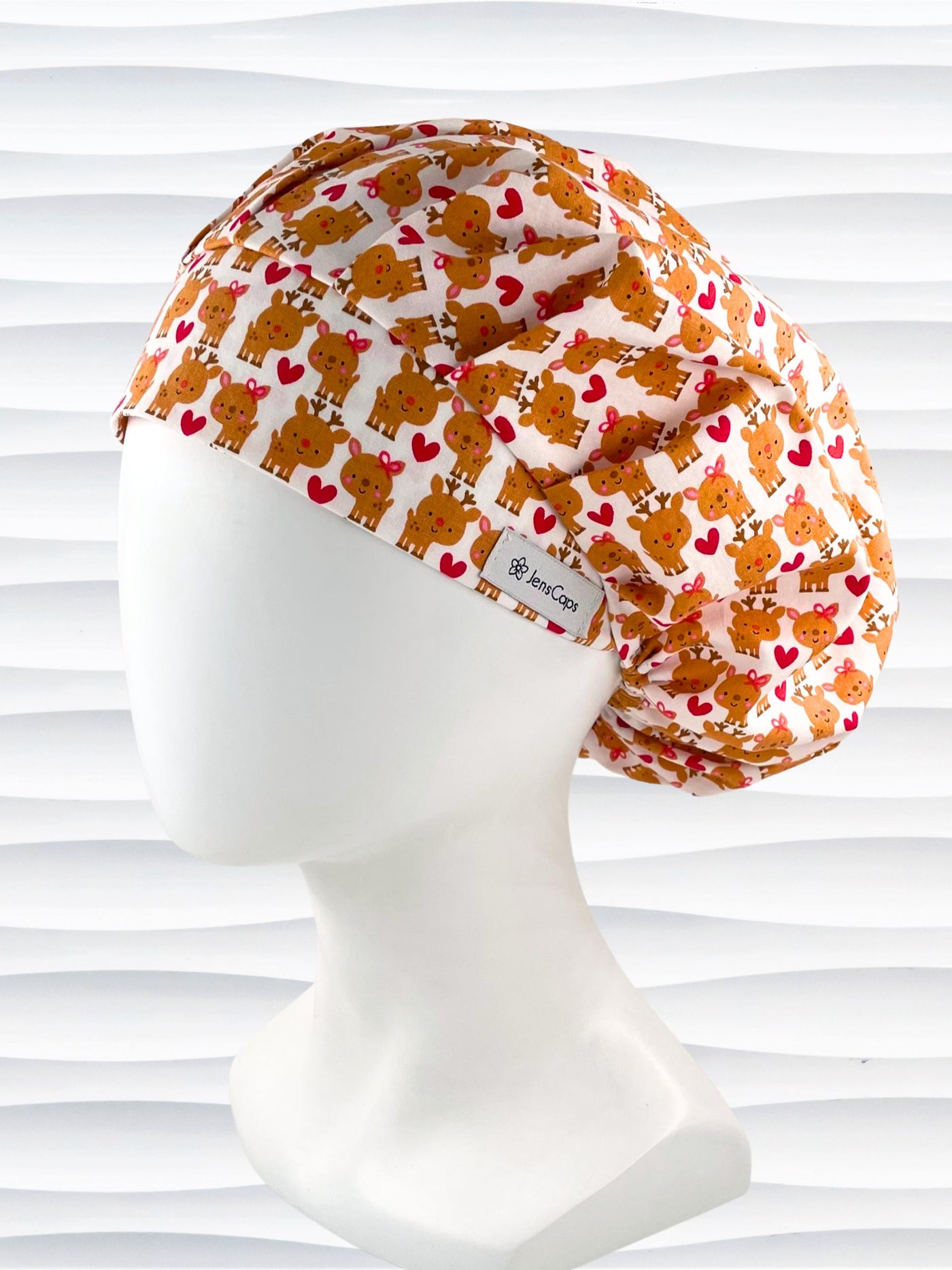 Bouffant style srugical scrub cap by Jenscaps made of white fabric patterned with repeating cute cartoon reindeer—some with pink bows—and red hearts, displayed against a white textured background.