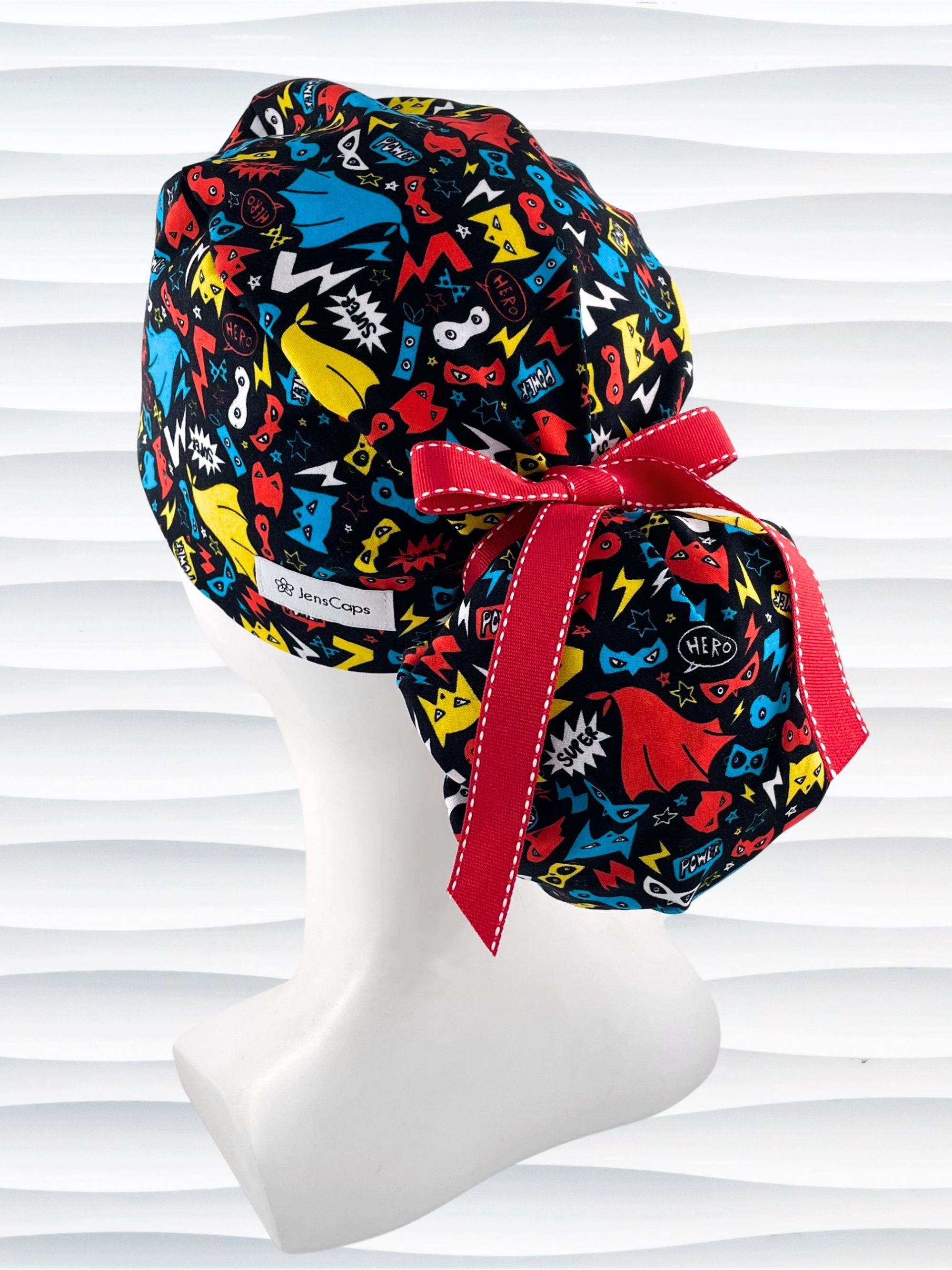 Ponytail style surgical scrub cap hat with superhero masks and capes in red, blue, and yellow on black cotton fabric.